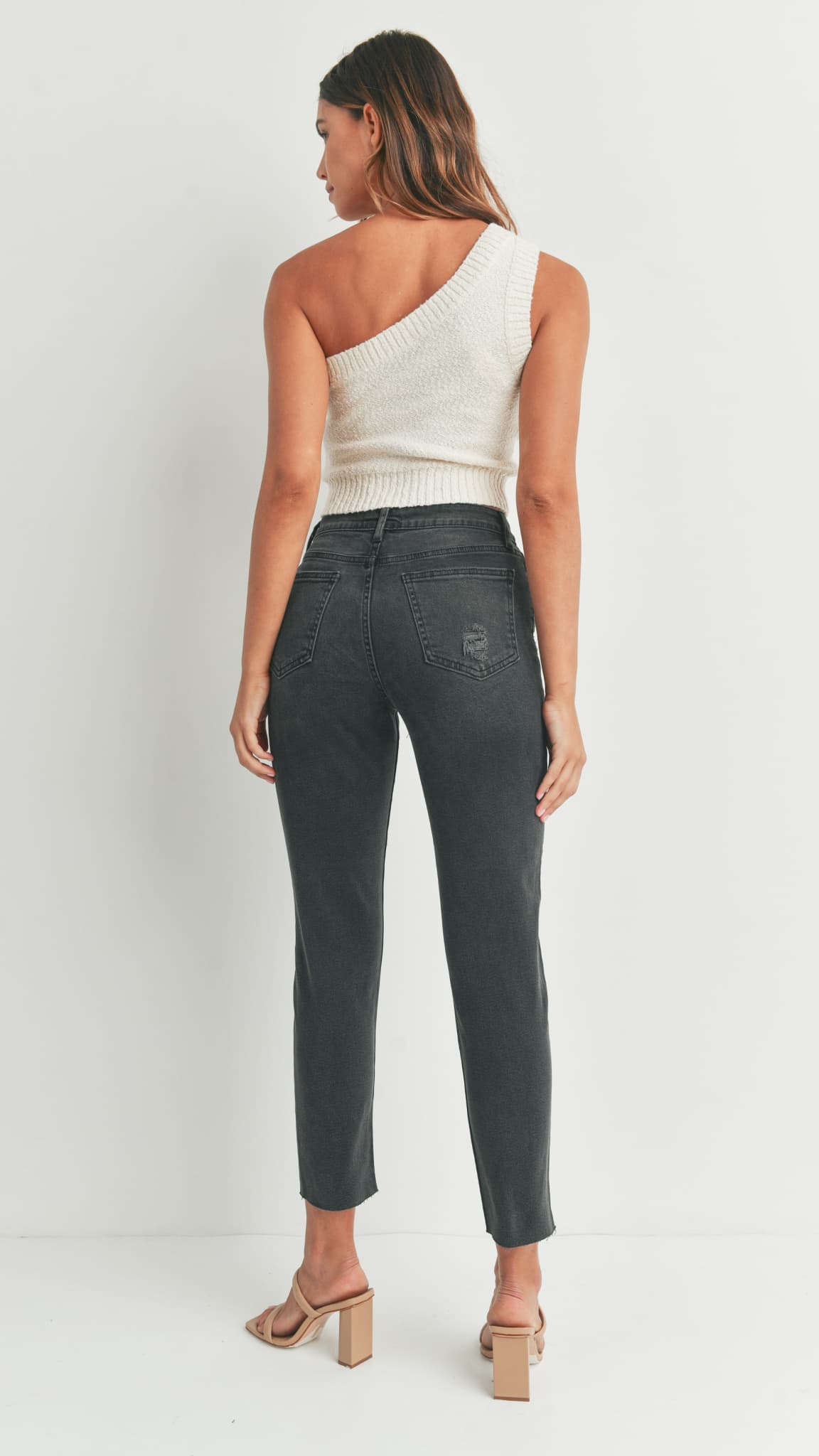 Back view of woman in black straight-leg jeans, white one-shoulder knit top, and beige heeled sandals