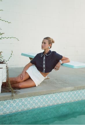 Woman lounging by a pool, wearing a navy short-sleeve collared top with white trim, and a white mini skirt