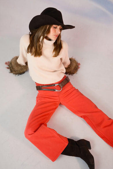 A woman in a snow setting is wearing a tan sweater, cowboy hat, black boots, and red jeans. She is sitting on the floor.
