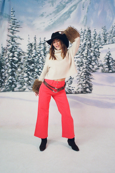 A woman in a snow setting is wearing a tan sweater, cowboy hat, black boots, and red jeans. She has a hand holding her cowboy hat on her head.