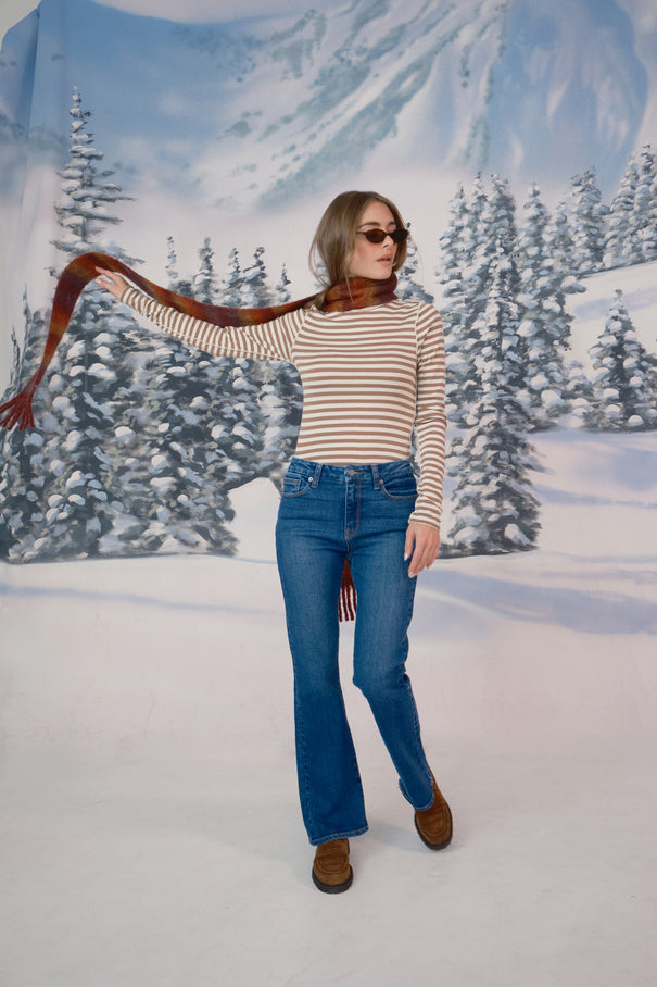 A woman in a snow setting is wearing a tan sweater, blue jeans, sunglasses, a scarf, and brown shoes. She is flailing her scarf to the side with one arm.