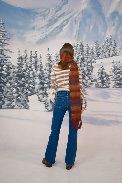 A woman in a snow setting is wearing a tan sweater, blue jeans, scarf, and brown shoes. She is turned with her back to the camera, standing straight. 