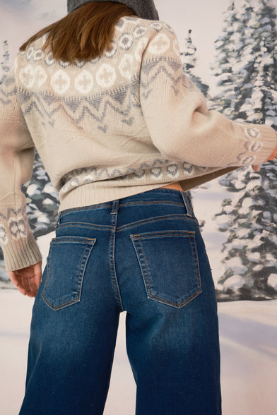 A woman in a snow setting is wearing a grey hood, a tan knit winter sweater, blue jeans. She has her back to the camera, tugging on her sweater. The show is close-up, showing primarily her shoulder to above the knee. 