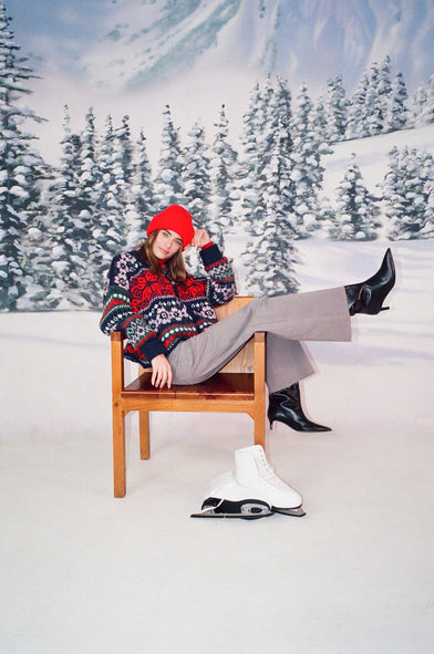 A woman in a snow setting is wearing a red beanie, knit winter sweater, and grey jeans. She is sitting in a chair with her leg kicked up into the air. There are ice skates on the floor. 