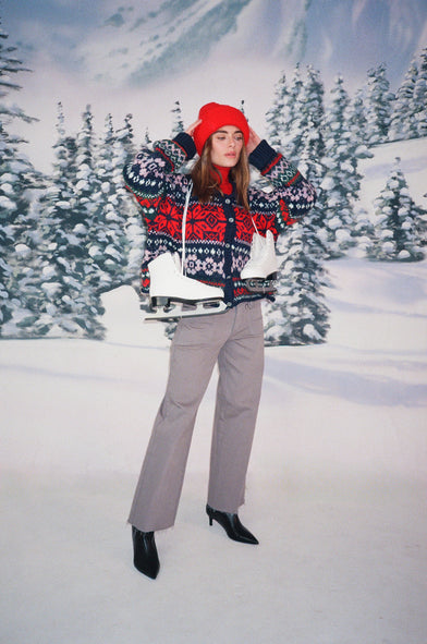 A woman in a snow setting is wearing a red beanie, knit winter sweater, and grey jeans. She has ice skates hung over her shoulders by the laces.