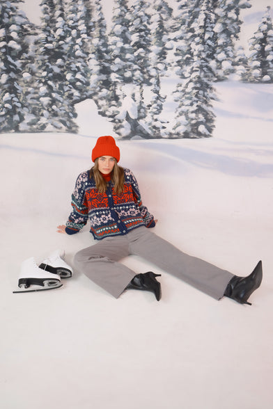 A woman in a snow setting is wearing a red beanie, knit winter sweater, and grey jeans. She is sitting in the snow with ice skates next to her. 