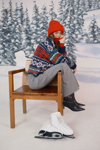 A woman in a snow setting is wearing a red beanie, knit winter sweater, and grey jeans. She is sitting in a chair with ice skates laying on the ground.