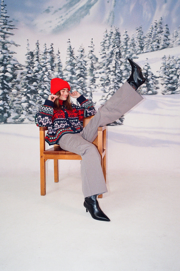 A woman in a snow setting is wearing a red beanie, knit winter sweater, and grey jeans. She is sitting in a chair with one leg kicking up into the air.