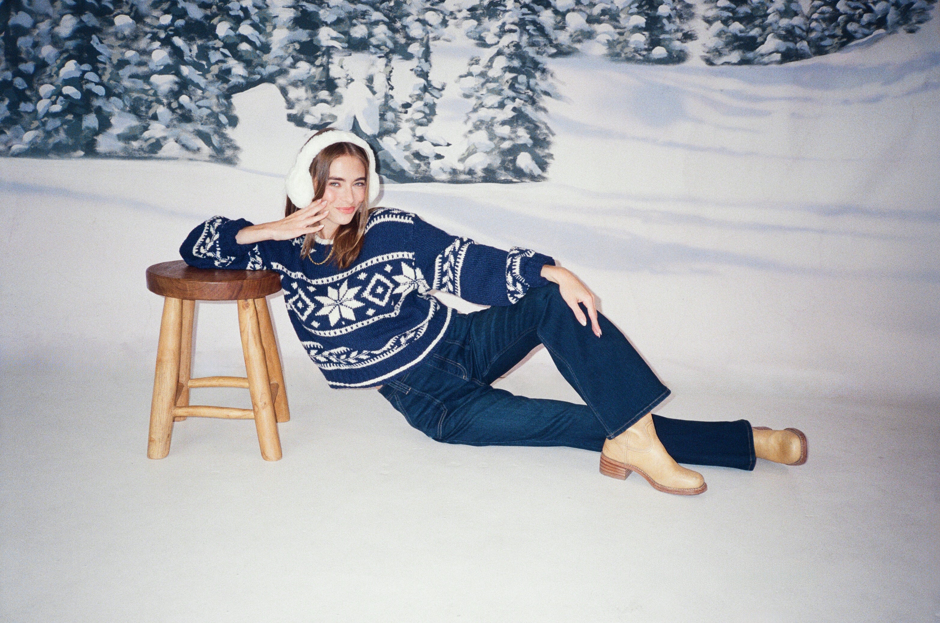 Woman is wearing white ear muffs, blue winter sweater, and blue jeans. She is laying in a snowy landscape with her arm resting on a stool. 