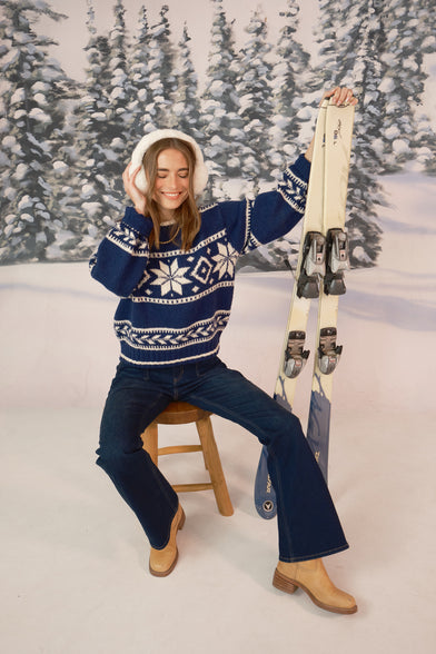 A woman in a snow setting is wearing a winter blue knit sweater, blue jeans, tan boots, and white fur ear muffs. She has one hand resting on her ear muff and the other hand is resting on her skis. She is sitting on a stool.