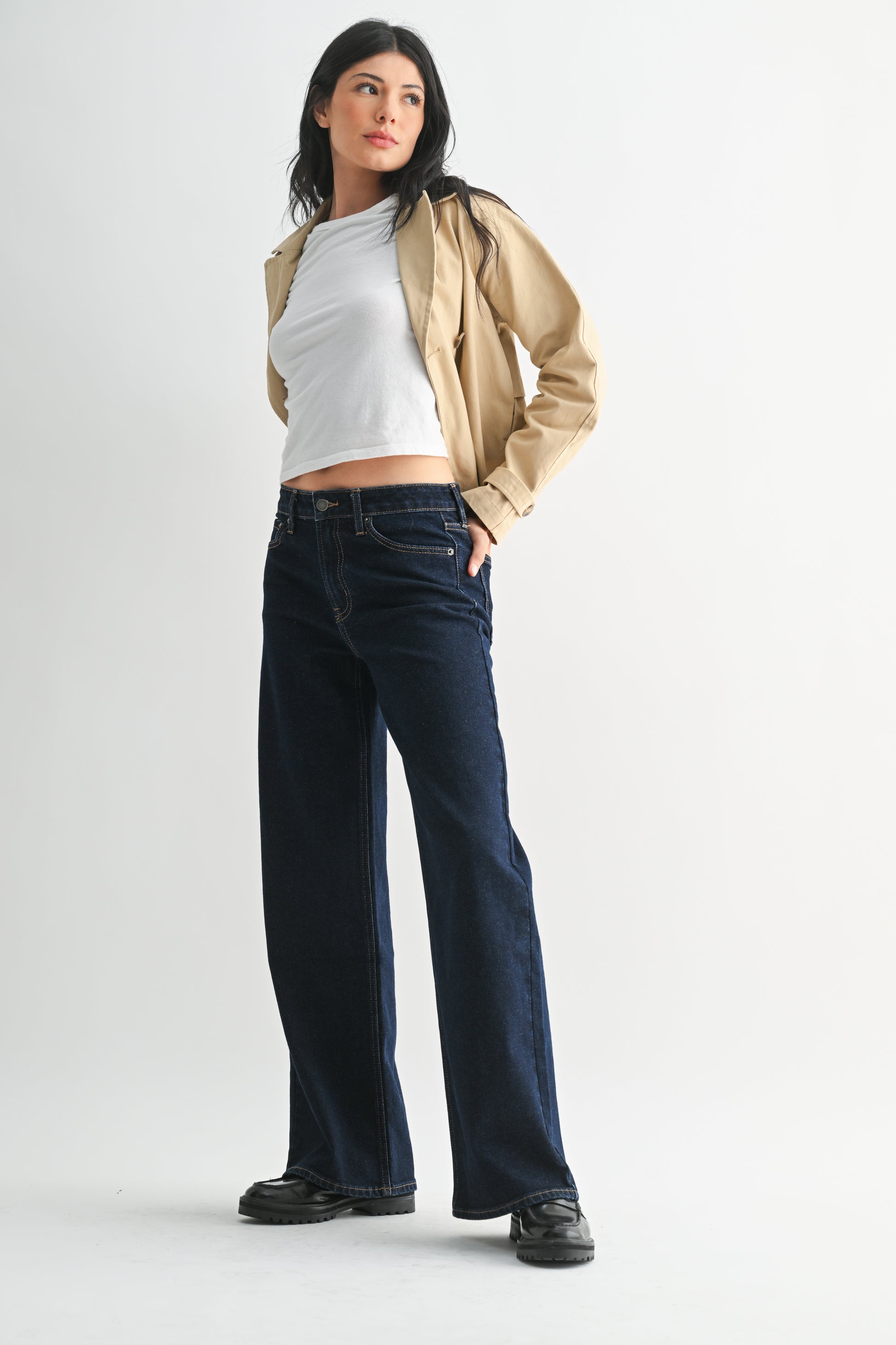 Woman wearing a beige jacket, white crop top, and dark blue jeans on a light gray background