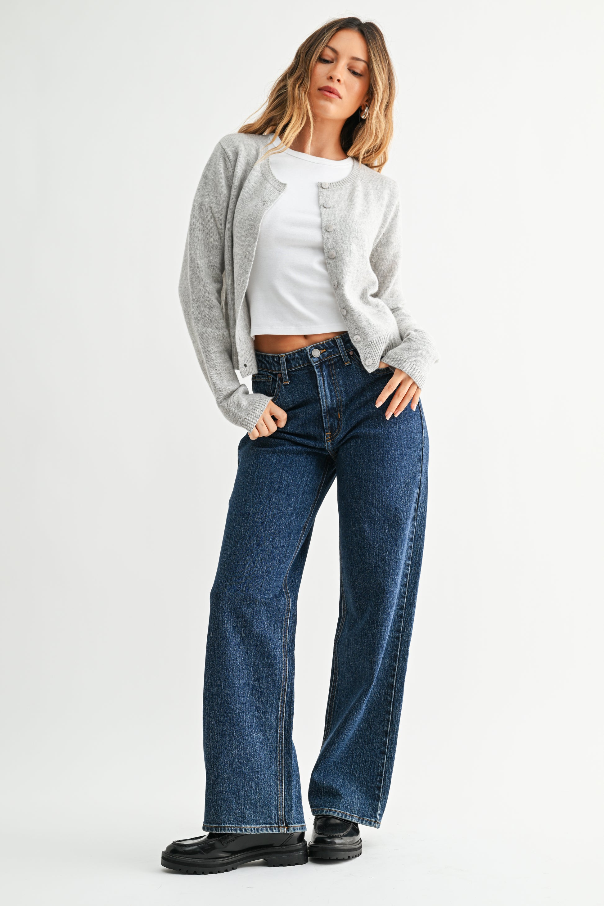 Woman wearing a gray cardigan, white top, blue jeans, and black shoes on a white background