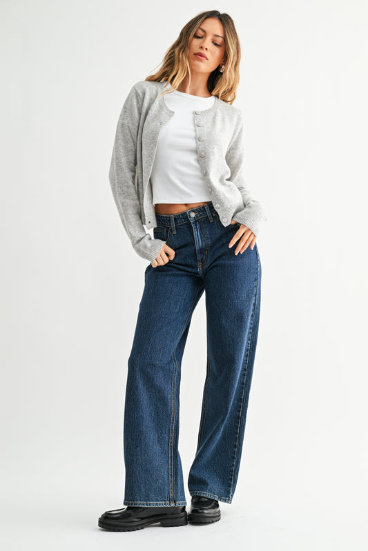 Woman wearing a gray cardigan, white top, blue jeans, and black shoes on a white background