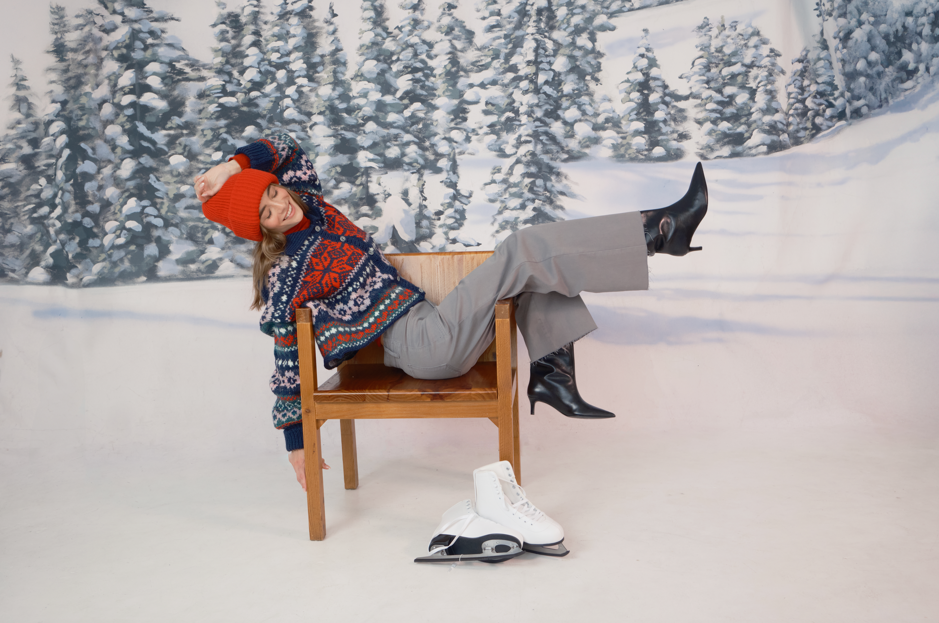 A woman lounging playfully on a wooden chair against a snowy forest backdrop. She is wearing a red knit beanie, a colorful patterned sweater, gray wide-leg pants, and black heeled boots. One arm dangles toward the floor, and her legs are lifted off the ground. A pair of white ice skates rests on the floor in front of the chair.